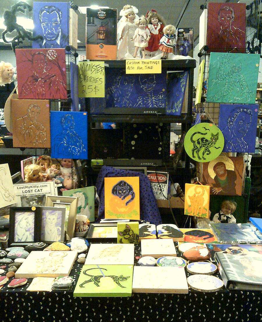 Table Display from Monster Bash 2026 My creepy horror and Halloween wares. Note the big, hairy spiders and creepy dollies peeping around the corners. Pieces at this show included blacklight art, lots of black cat art and classic horror-genre actor paintings.
